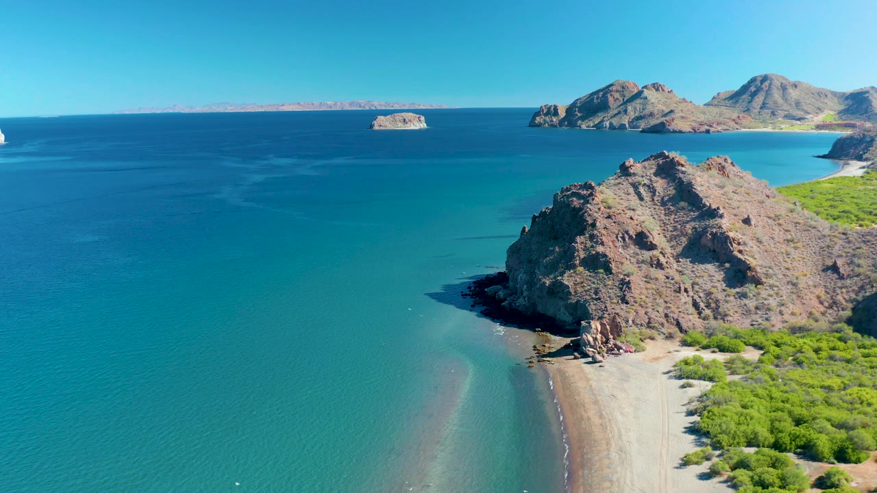 At 0:02 the aerial view over Ligui shows the rocky coast slightly farther right, calm layered blue water across most of the frame, distant islands near the horizon, and no visible people.
