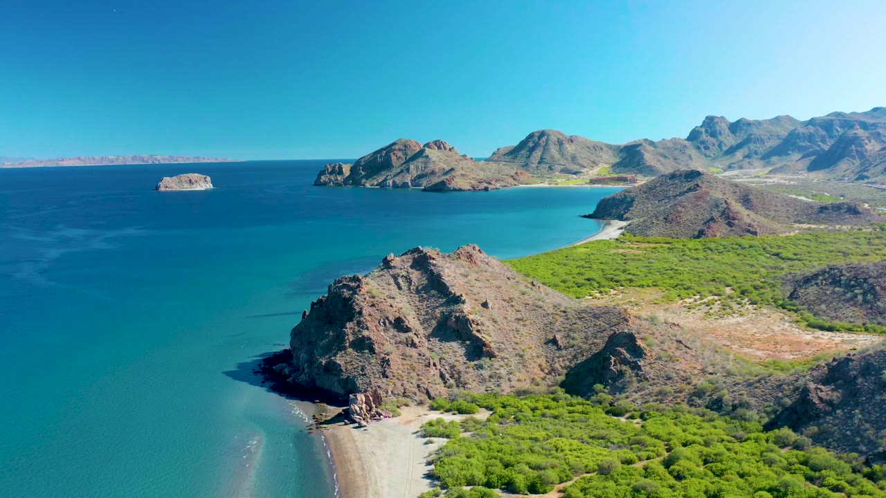 Aerial frame from Ligui, Baja California Sur, showing the foreground cliff shifted slightly lower in view, with the sandy beach edge, turquoise water, offshore rocks, and no visible people.