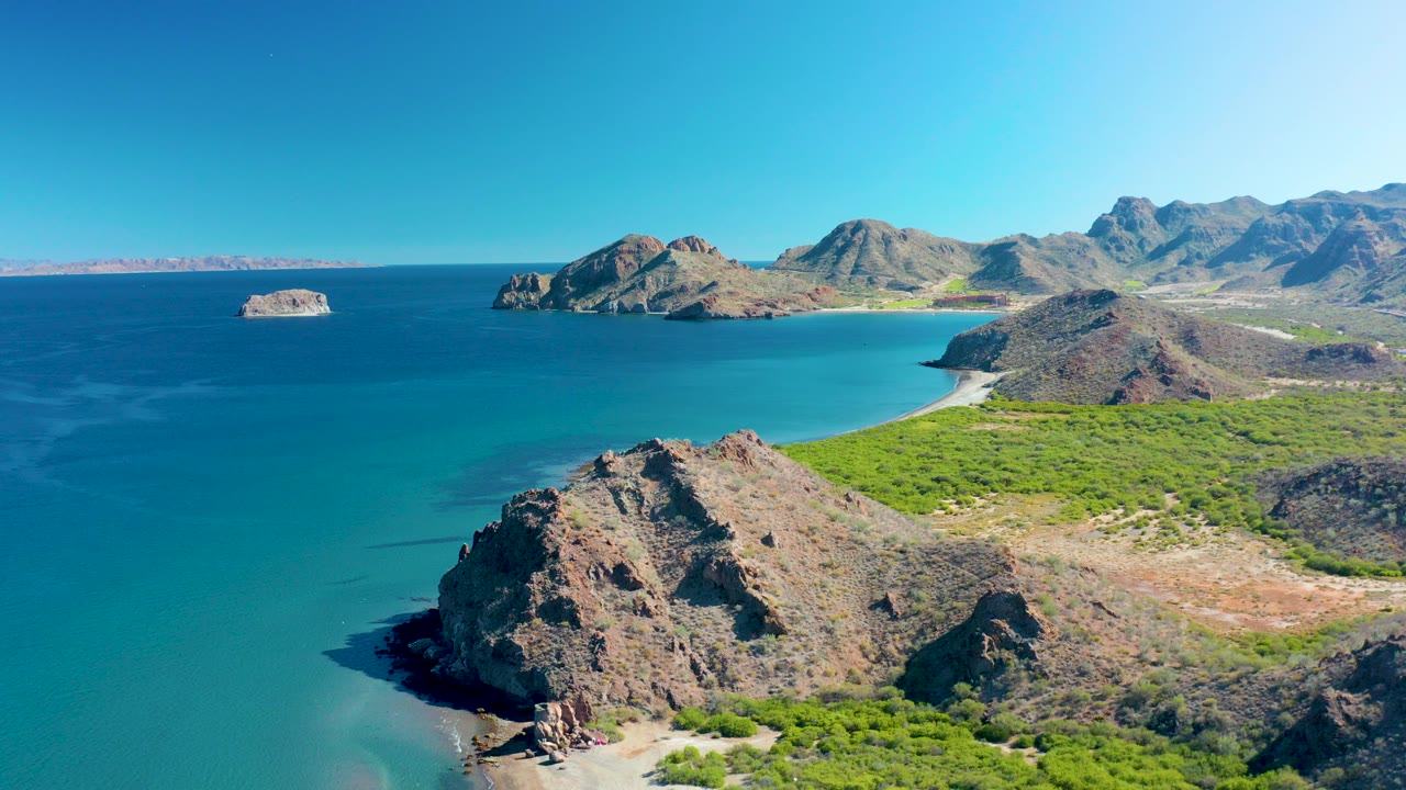 Aerial view over Ligui, Baja California Sur with a rocky foreground headland, turquoise bay water, green shore vegetation, distant islands, mountain ridges, and no visible people.