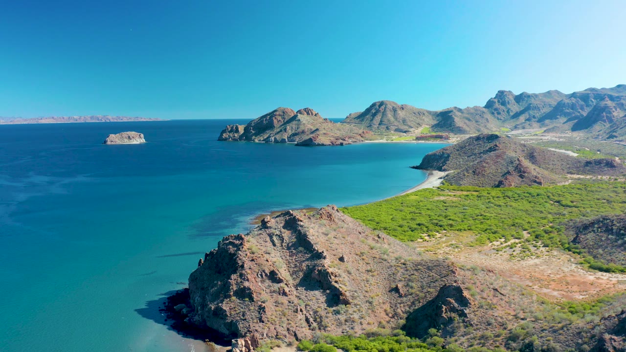 Wide aerial bay with rocky point, green coastal plain, blue ocean, and mountains.