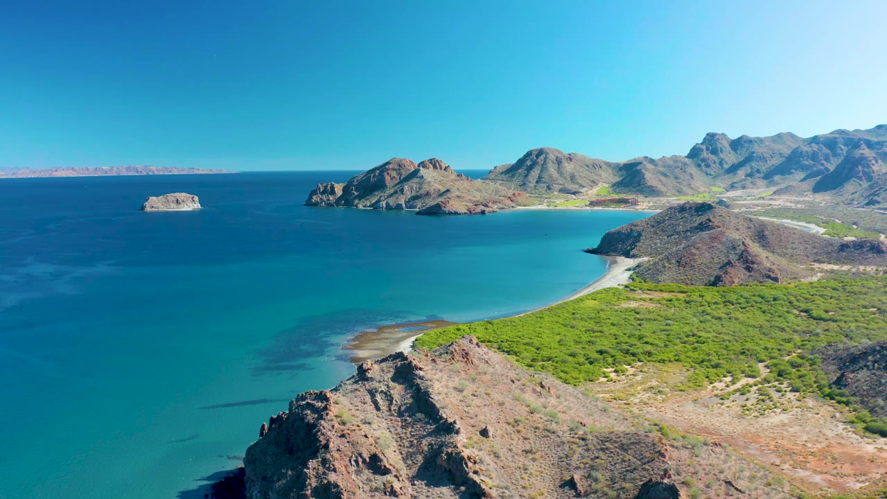 Aerial frame in Ligui, Baja California Sur showing the bay filling the left side, a pale beach at center, rocky headlands and mountains beyond, and no visible people.