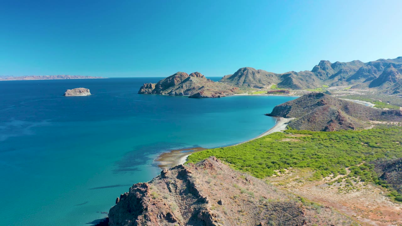 High aerial view of Ligui coastline with shallow turquoise water near the sand, darker blue ocean farther out, rocky desert hills, distant mountains, and no visible people.