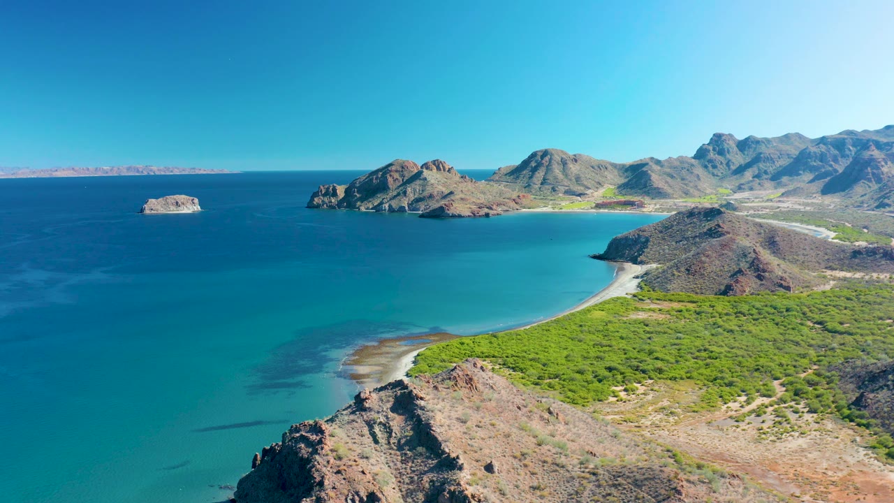 Aerial view across calm blue water toward beach, cliffs, islands, and mountains.