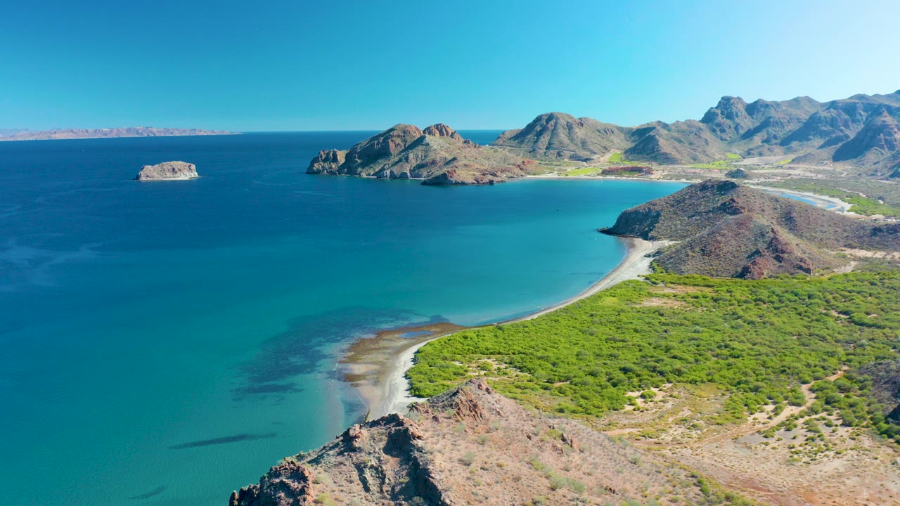 Wide aerial frame over Ligui, Baja California Sur showing turquoise shallows around a crescent beach, rocky hills, distant mountains, a small offshore rock, and no visible people.