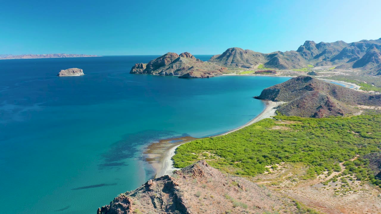 Aerial view in Ligui, Baja California Sur with the curved sandy shoreline lower in frame, green vegetation across the beach edge, rocky headlands beyond, and no visible people.