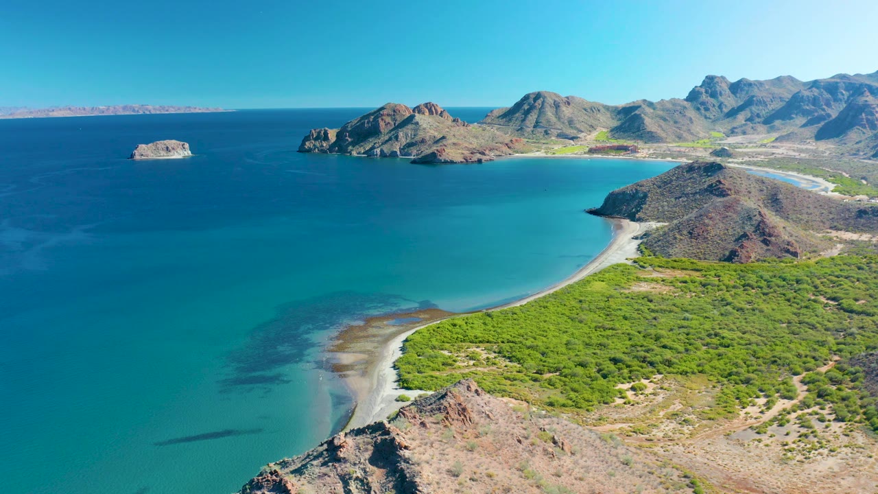 Wide aerial beach view with offshore rock, turquoise water, and rugged hills.