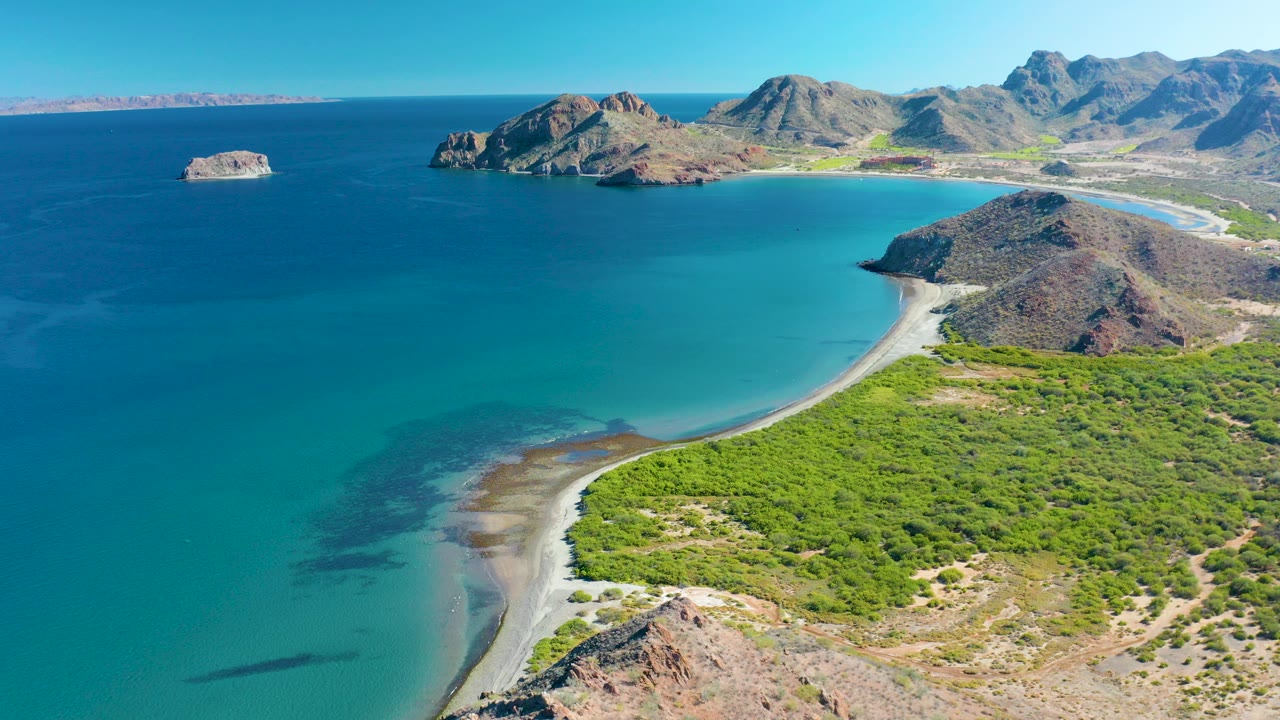 Aerial view over Ligui, Baja California Sur, with a sandy beach curving around turquoise water, green vegetation in the foreground, rocky hills in back, and no visible people.