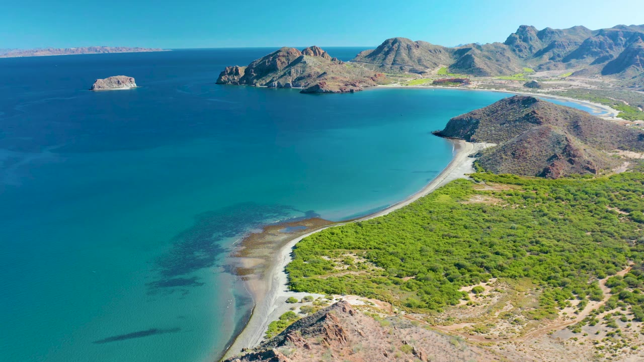 Wide aerial frame in Ligui showing the turquoise bay filling the center, the pale sandy shoreline bending along green brush, rocky mountains beyond, and no visible people.