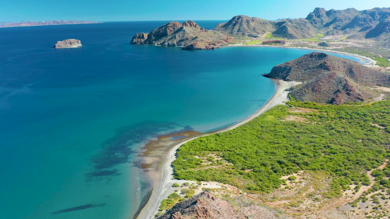 High coastal view of turquoise bay water, pale sand, green shoreline, and distant rocky terrain.