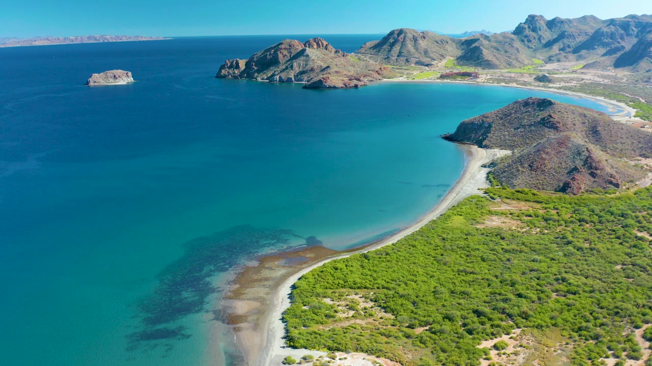 Aerial frame over Ligui, Baja California Sur, showing a turquoise bay curving around a sandy shore, green coastal scrub in front, rocky islands offshore, mountain ridges in the distance, and no visible people.