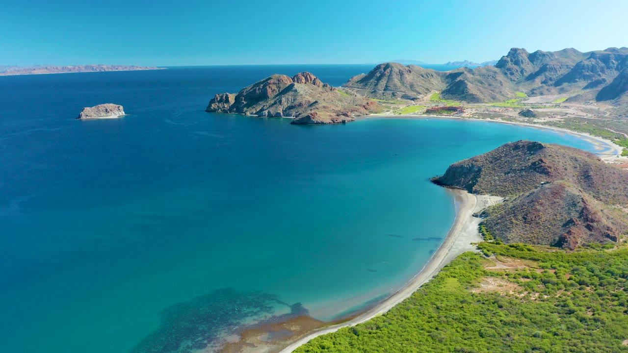 Wide aerial view over Ligui, Baja California Sur showing a blue bay, curved sandy beach, rocky hills, green shoreline vegetation, distant mountains, and no visible people.