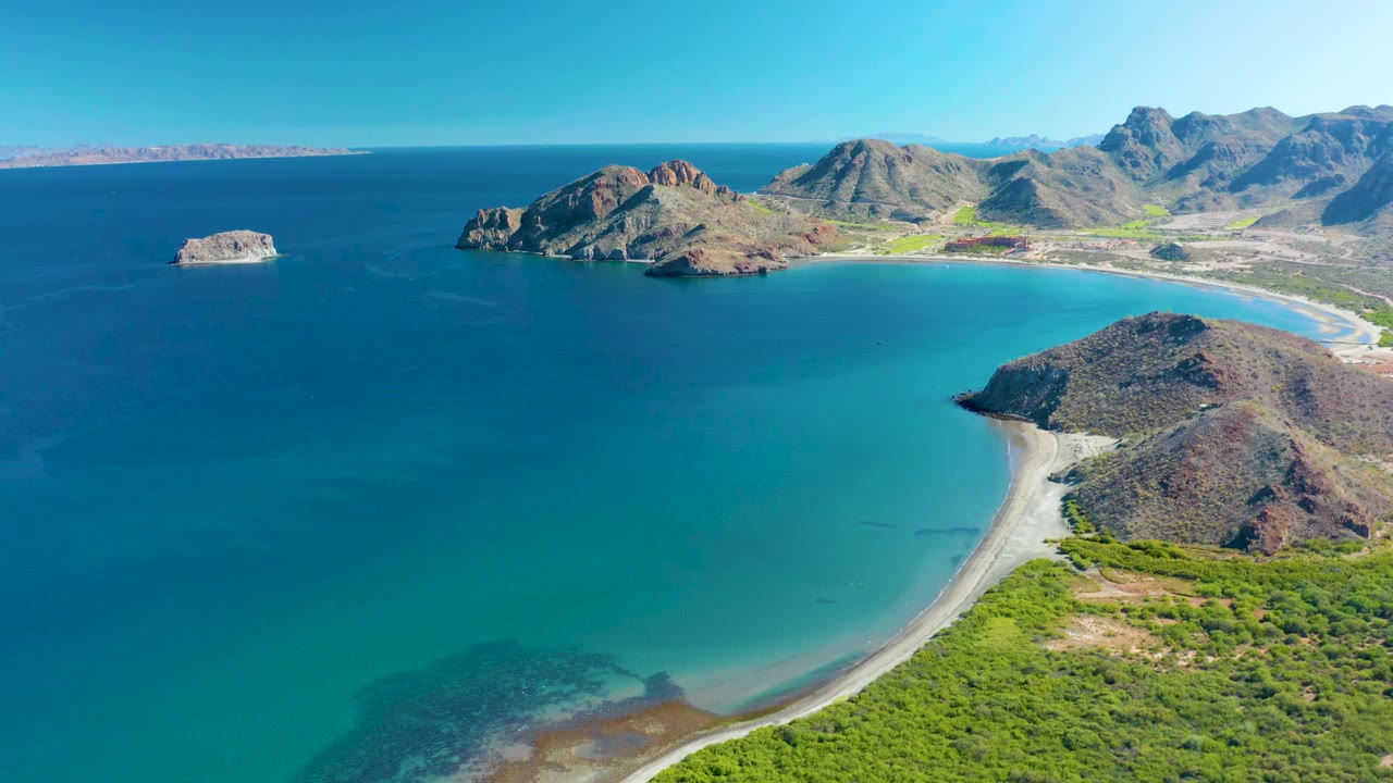 Aerial frame over Ligui with more open water centered in the bay, the pale curved beach along the right edge, rocky hills behind it, a small offshore rock, and no visible people.