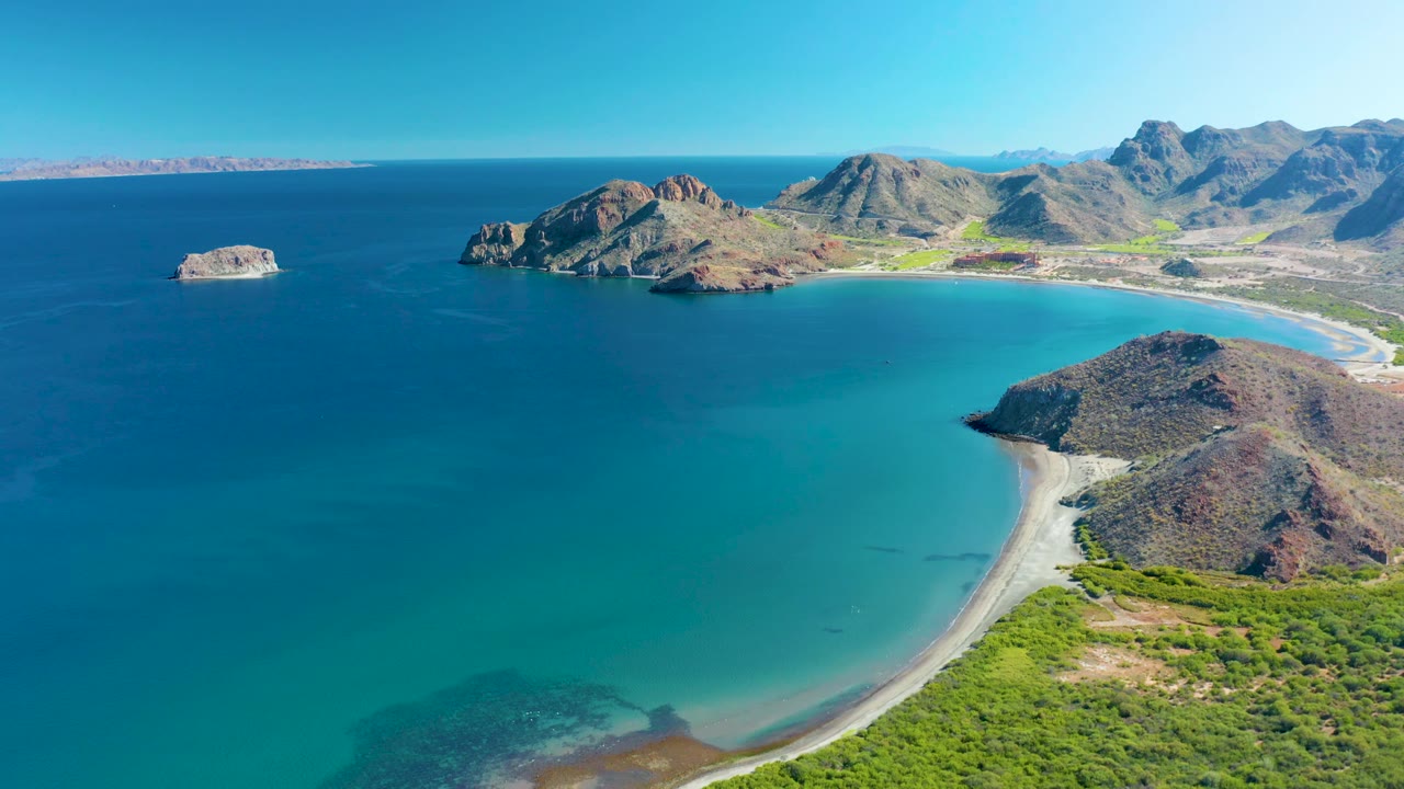 A high coastal view of Ligui shows the rocky point lower right, the sandy arc of beach behind it, deep blue water to the left, distant mountain ridges, and no visible people.
