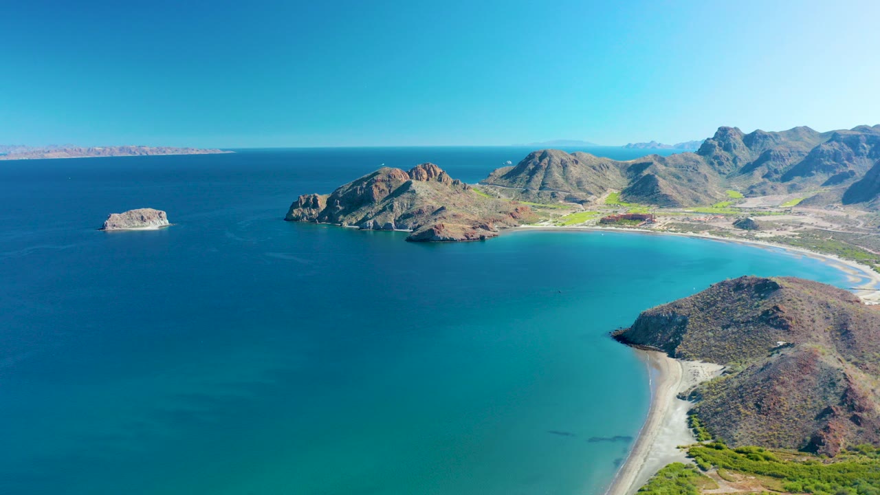 Aerial view over Ligui, Baja California Sur, showing turquoise water curving into a sandy shore, rocky headlands, distant mountains, and no visible people.
