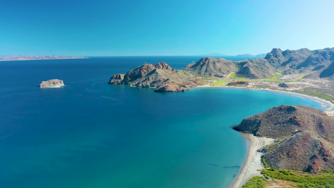 Wide aerial frame in Ligui with deep blue ocean on the left, a small offshore rock, a turquoise bay, sandy beach, mountain ridges, and no visible people.