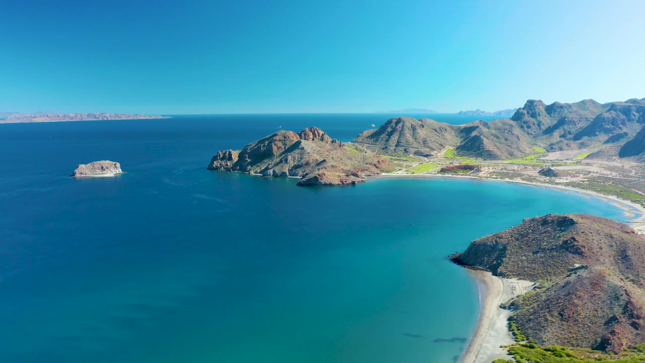 High aerial frame over Ligui showing the offshore rock at left, the main rocky headland near center, a sandy shoreline to the right, mountain ridges behind, and no visible people.