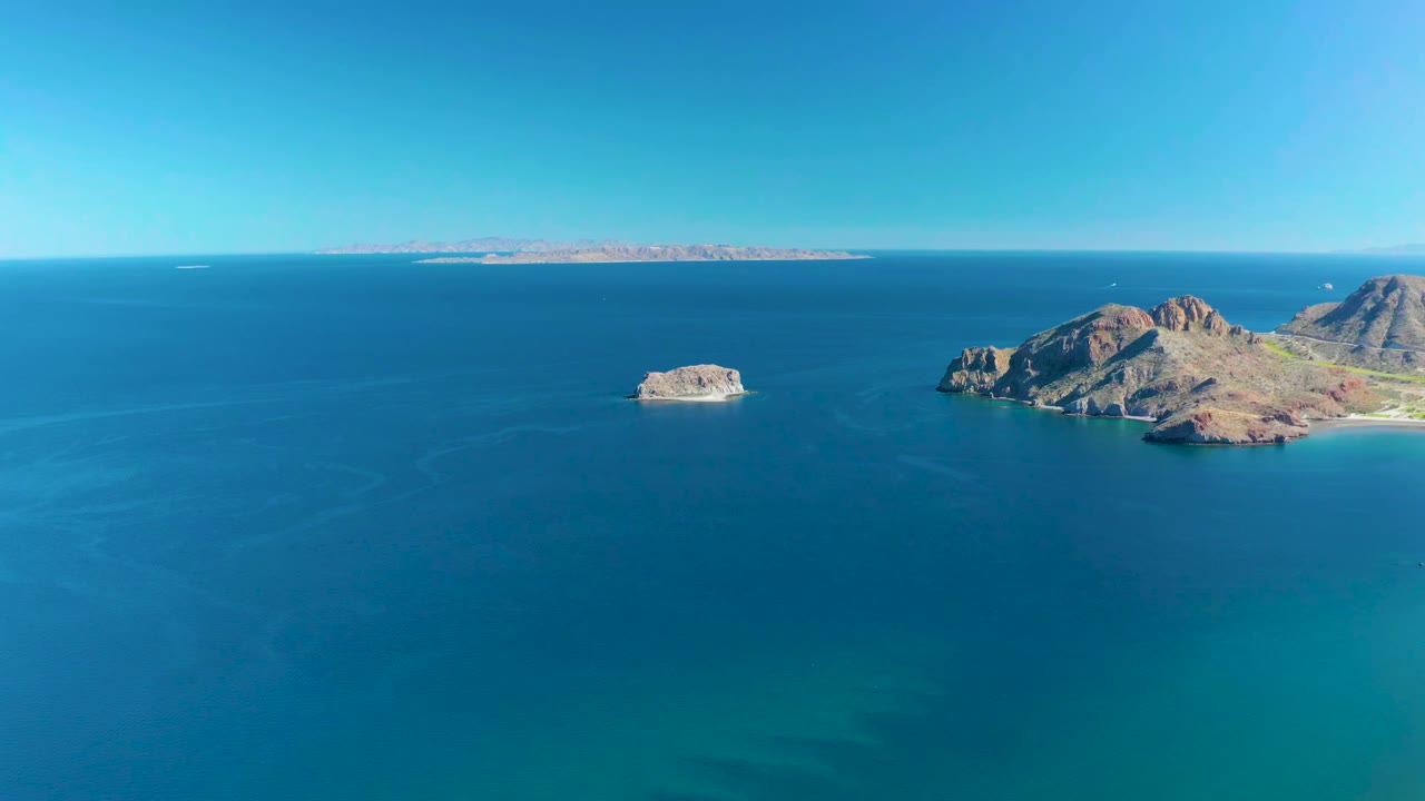 Wide aerial frame over Ligui, Baja California Sur, showing blue ocean, a small offshore rock, larger rocky islands to the right, distant coastline on the horizon, and no visible people.