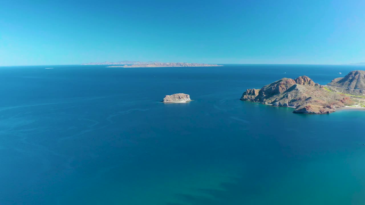 Aerial view at 0:01 over Ligui with the small rock left of center, rugged islands on the right edge, calm blue water across the foreground, and no visible people.
