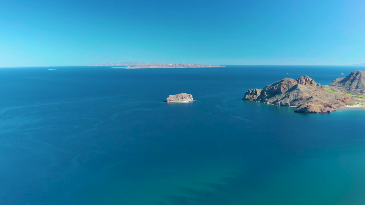 Aerial view in Ligui, Baja California Sur, with the small white boat slightly farther across the blue water, rocky islands on the right, distant shoreline on the horizon, and no visible people.