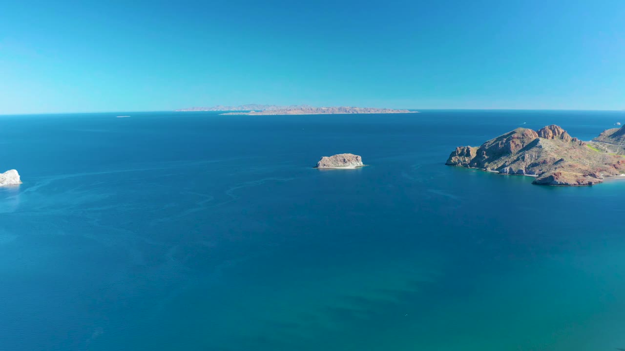 Aerial view in Ligui, Baja California Sur, Mexico, showing blue water across the foreground, a small rock island left of center, the larger rocky island to the right, pale distant coastline, and no visible people.