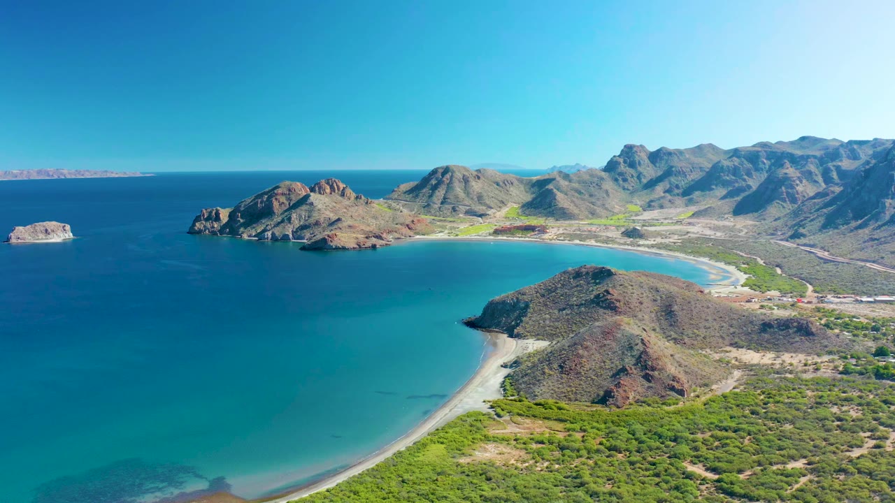 Aerial view over Ligui with the bay filling the left side, a pale beach wrapping the foreground hill, rugged mountains behind, and no visible people.
