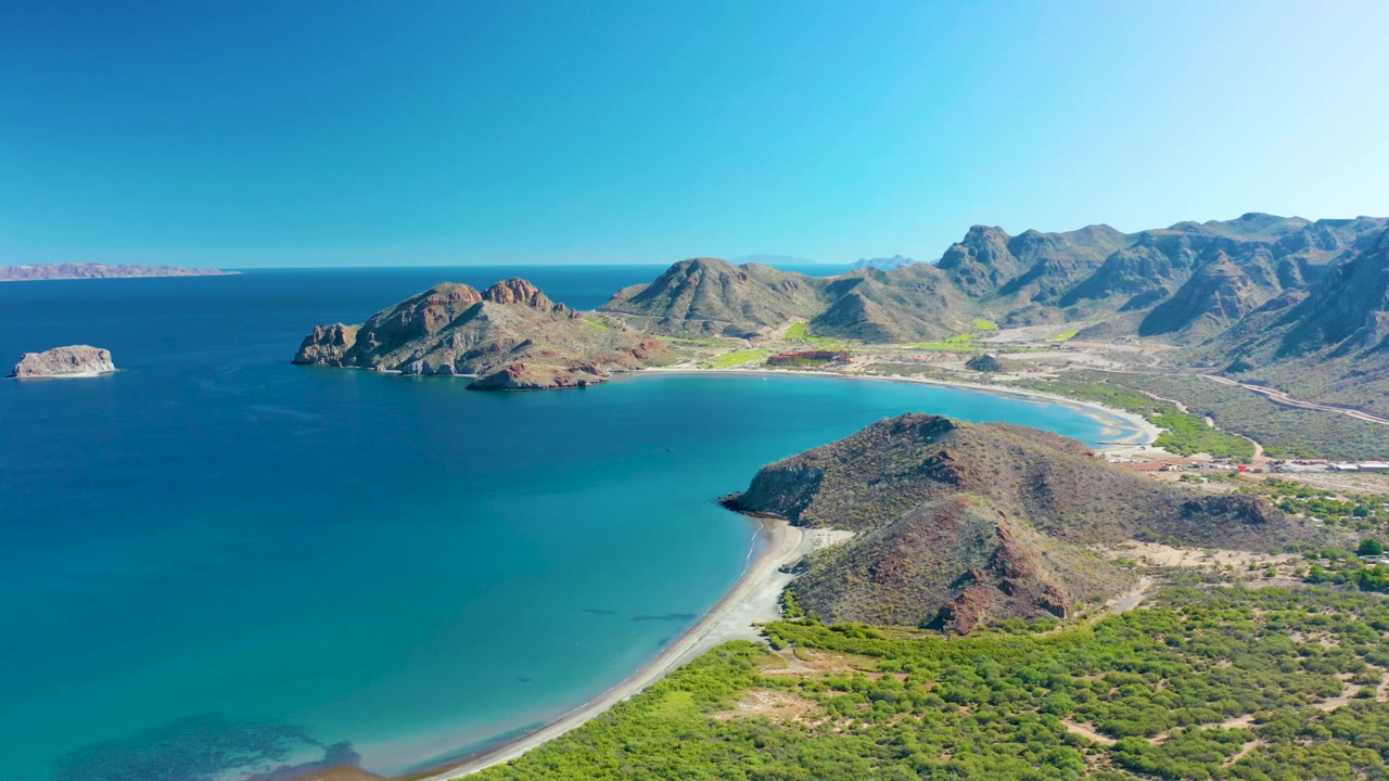 Drone frame from Ligui, Baja California Sur, with turquoise water curving around a sandy shoreline, rocky coastal hills to the right, a small offshore rock to the left, and no visible people.