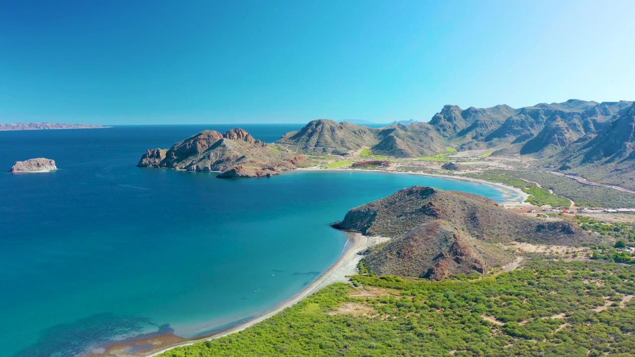 Opening aerial view over Ligui, Baja California Sur with turquoise water, a curved sandy beach, rocky headlands, mountain ridges, and no visible people.