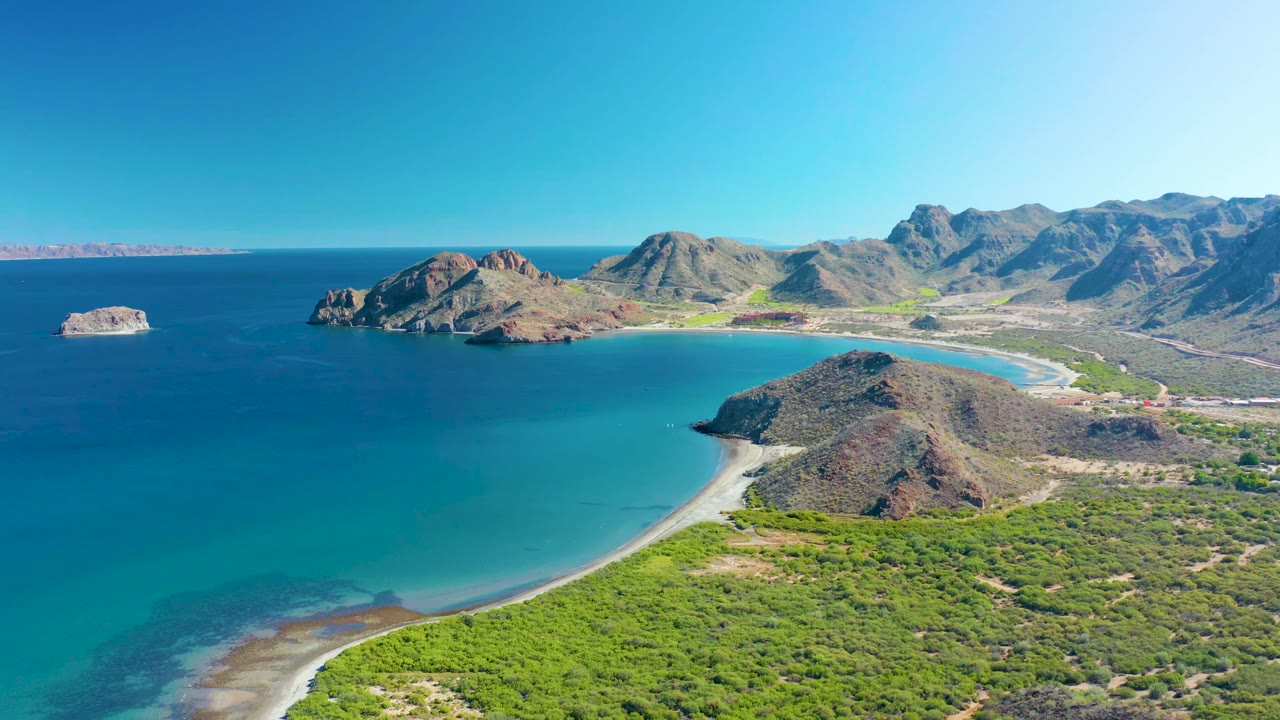 Wide aerial frame over Ligui showing a small offshore rock at left, deep blue ocean, a curved turquoise bay, sandy beach, green coastal vegetation, rocky hills, and no visible people.