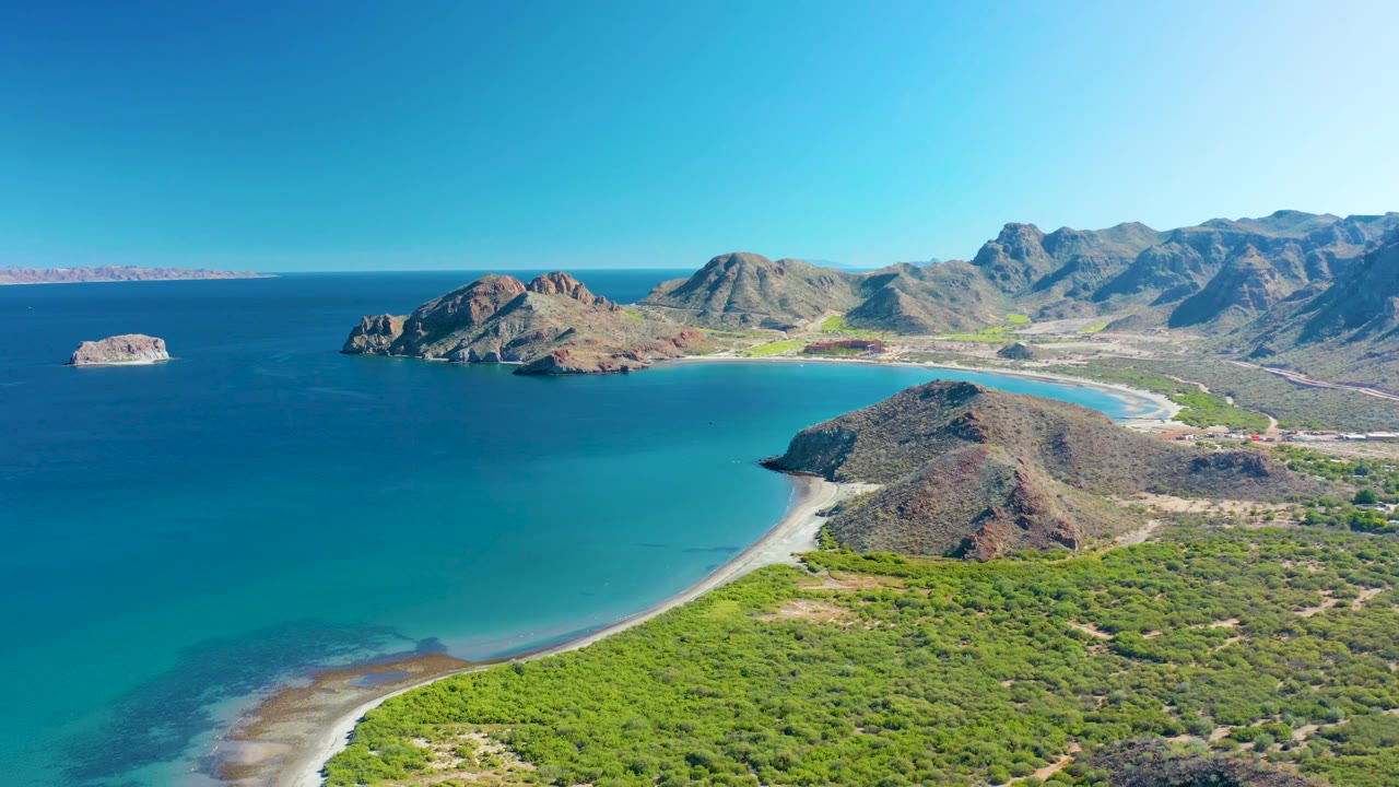Aerial view of Ligui's curved shoreline with pale sand, turquoise shallows, a rocky headland near center frame, green scrub below, distant hills to the right, and no visible people.