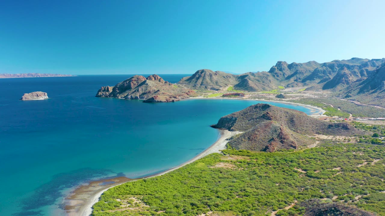 High aerial view over Ligui with broad blue ocean at left, a bright sandy beach wrapping the bay, green vegetation in the foreground, rugged hills along the right side, and no visible people.