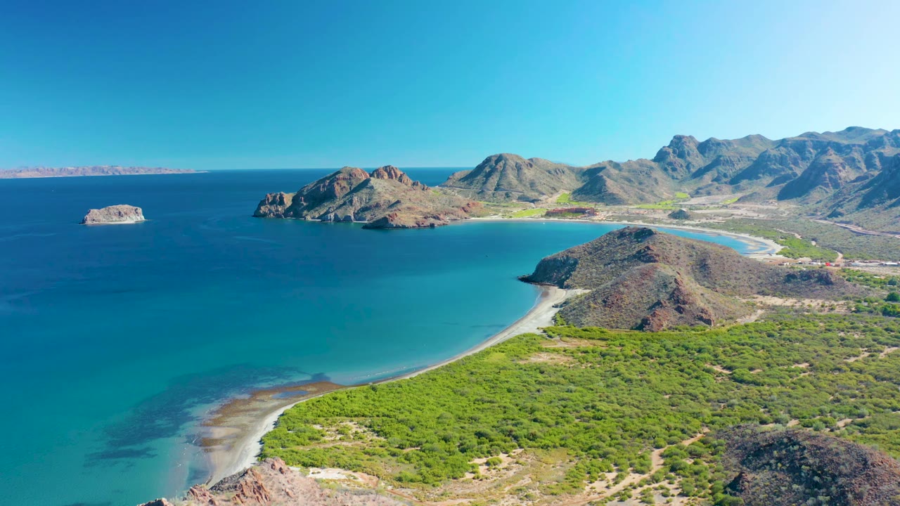 Aerial frame over Ligui with the sandy beach arc slightly lower in view, blue-green bay water, rocky outcrops, mountain ridges, clear sky, and no visible people.