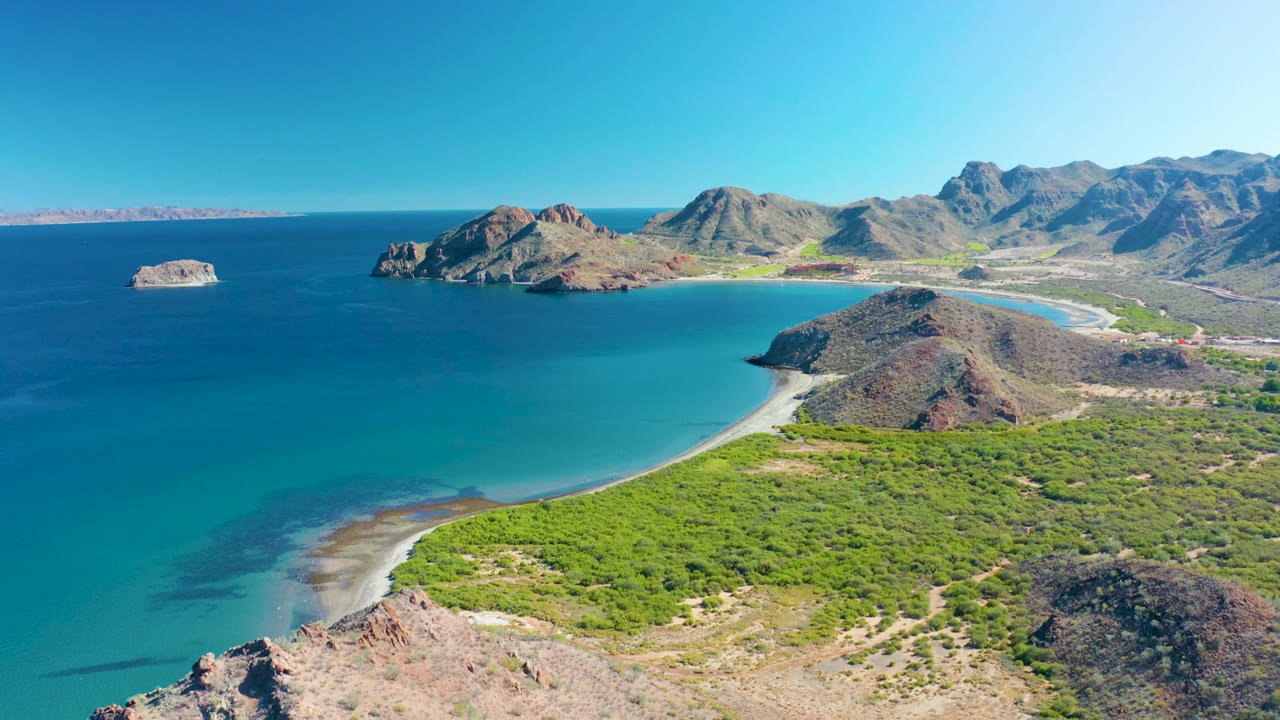 Mid-clip aerial over Ligui showing more rocky terrain at the bottom edge, a turquoise cove, broad green shoreline scrub, headlands, distant mountains, and no visible people.