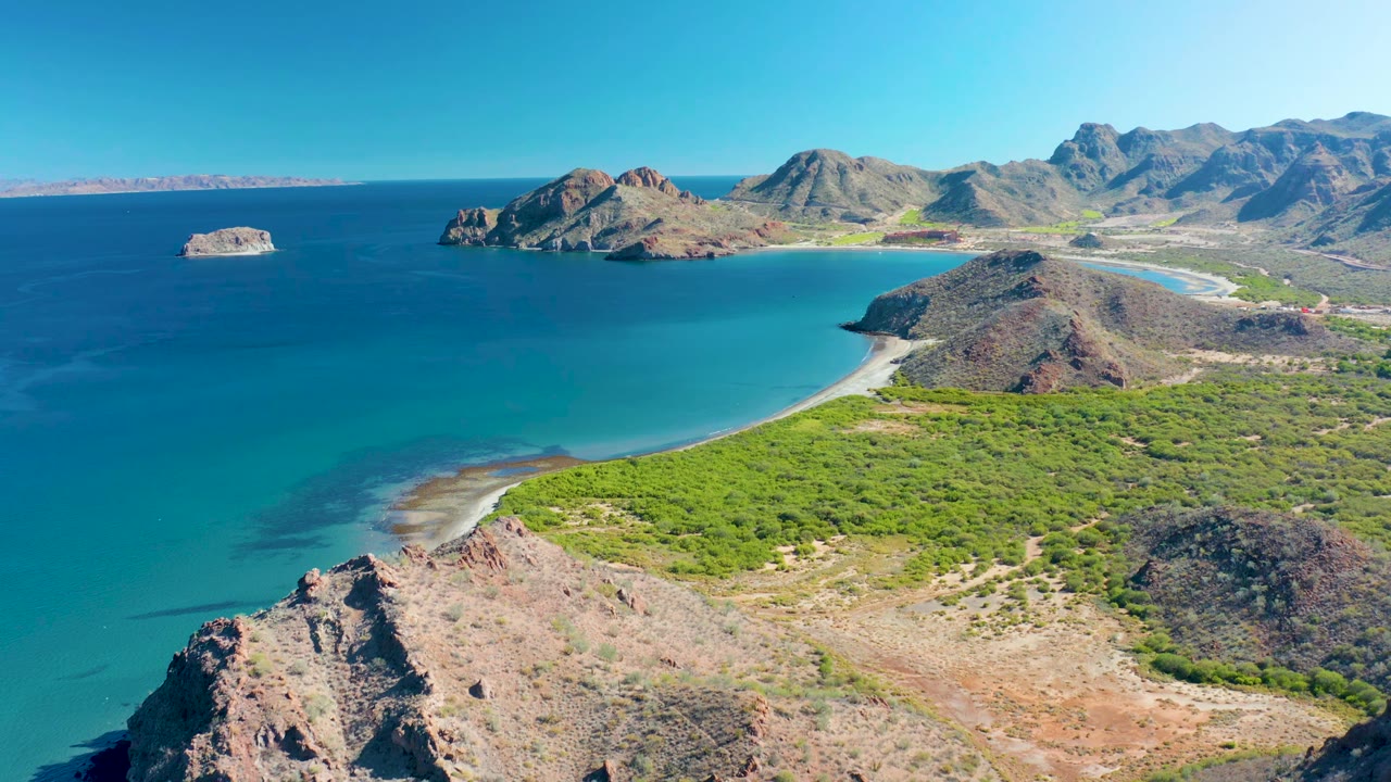 Wide aerial frame over Ligui coastline with the small offshore island left of center, a pale beach wrapping the bay, rocky headlands in the foreground and distance, and no visible people.