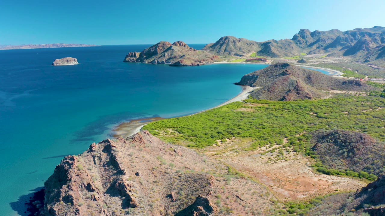 Aerial coastal landscape in Ligui, Baja California Sur, with a rocky foreground ridge, calm blue water, sandy beach, low green vegetation, mountain terrain, and no visible people.
