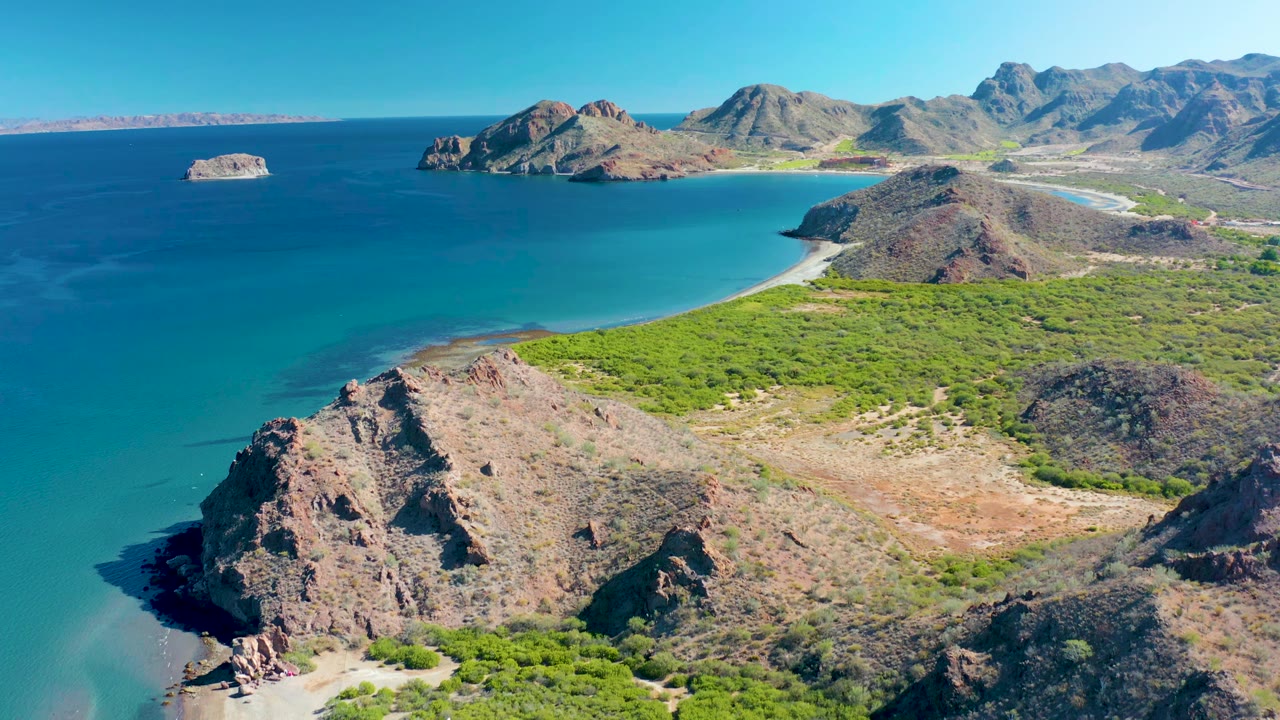 A high drone-style frame over Ligui shows the rocky foreground pushed closer to the beach curve, with green vegetation, blue bay water, distant rocky islands, mountains, and no visible people.