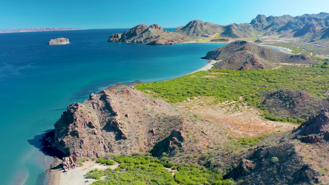 The aerial view over Ligui frames a broad rocky slope above the sand, a bright green coastal strip, turquoise water, distant islands on the horizon, and no visible people.