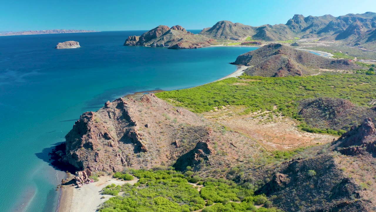 Mid-clip aerial frame over Ligui with the sandy beach entering at lower left, a central rocky hill, dense green brush, wide turquoise bay, distant mountains, and no visible people.