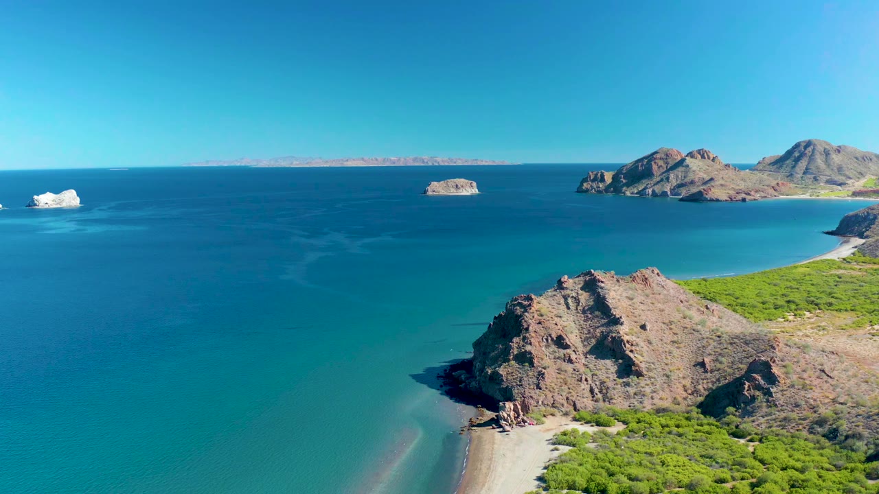 Wide aerial frame of Ligui coastline with the rocky point lower right, green vegetation along the shore, deep blue water beyond, small islands in the distance, and no visible people.
