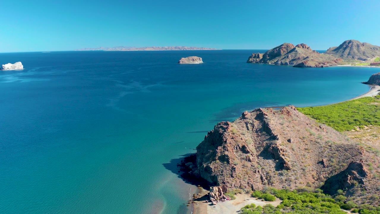 Aerial frame over Ligui, Baja California Sur with a rocky foreground headland, turquoise shallows, deep blue ocean, distant islands, and no visible people.