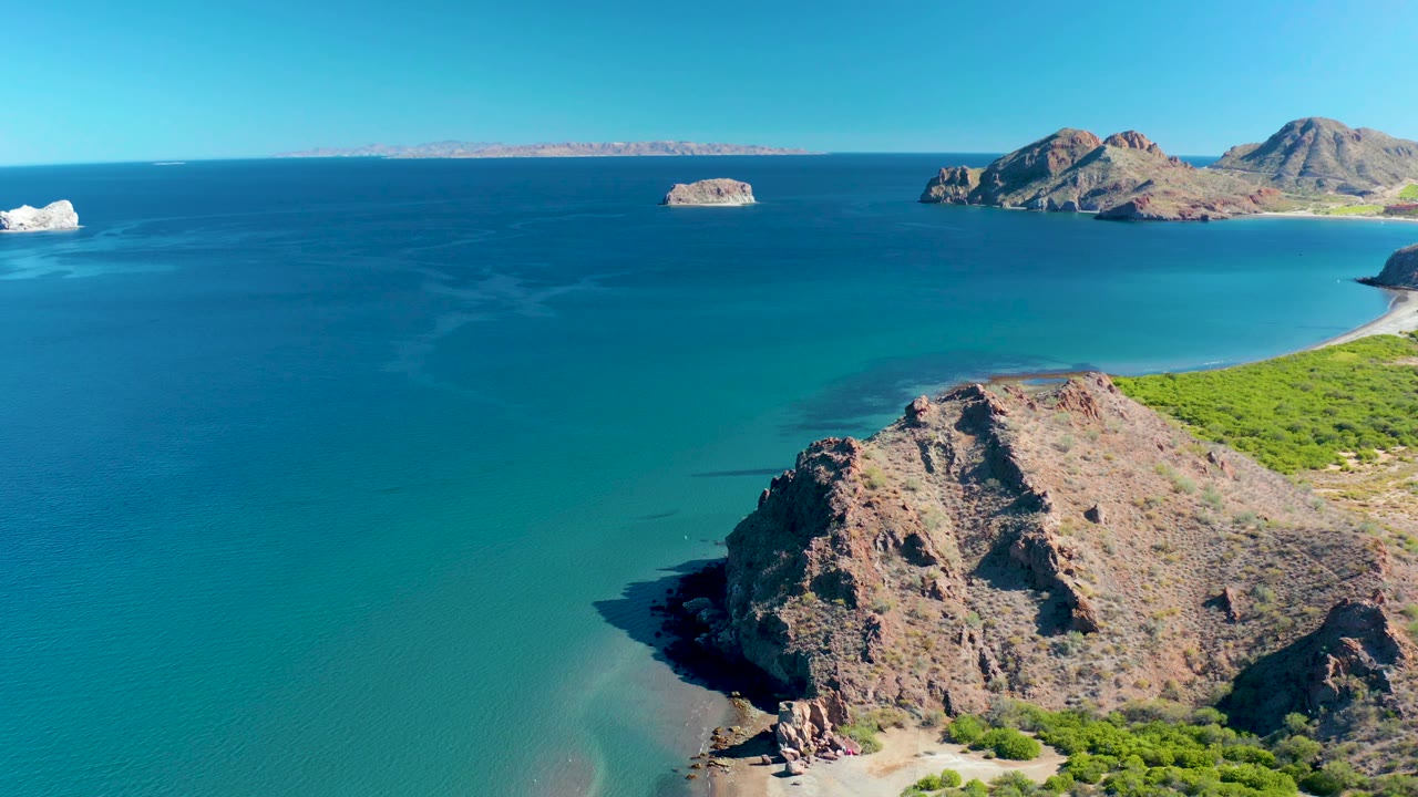 Ligui coastline frame with the rocky cliff mass low in the image, green shoreline to the right, calm blue ocean across the center, distant islands, and no visible people.