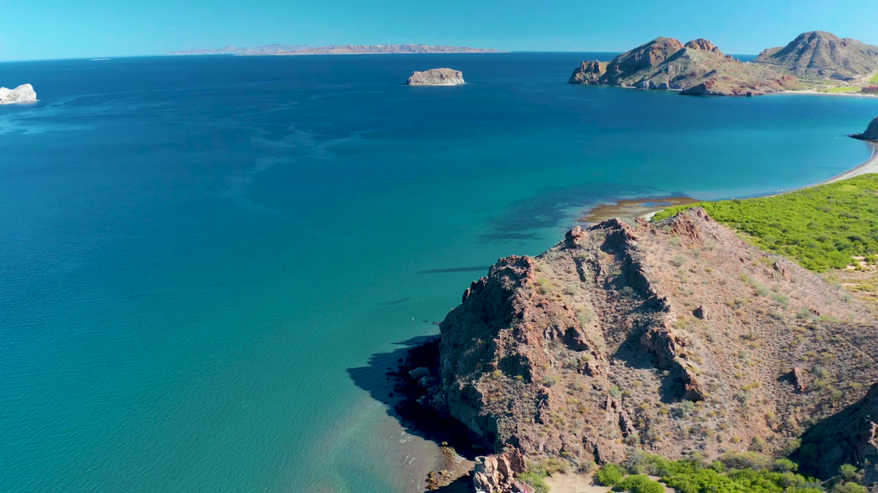 Aerial frame in Ligui showing the foreground rock face slightly closer, calm blue-green water below, island forms on the horizon, sparse vegetation, and no visible people.
