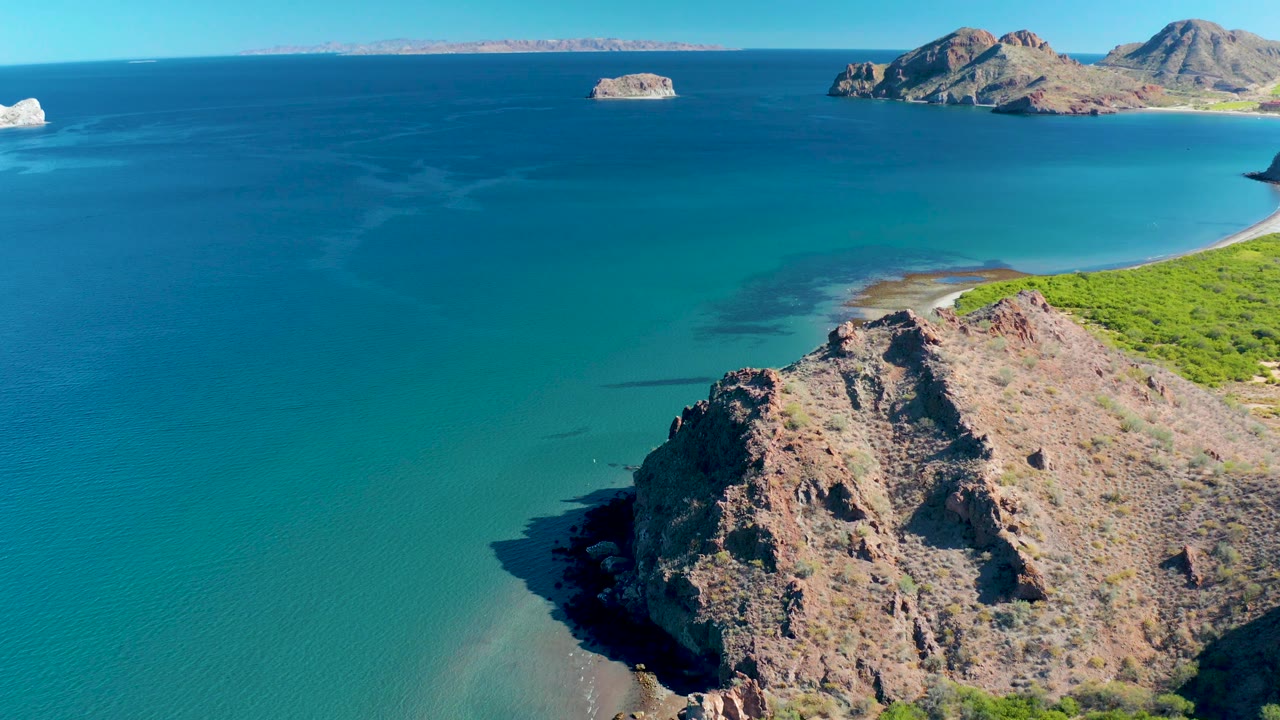 Mid-clip aerial view over Ligui with the rocky point shifting lower in frame, green vegetation along the right shoreline, still water, distant islands, and no visible people.