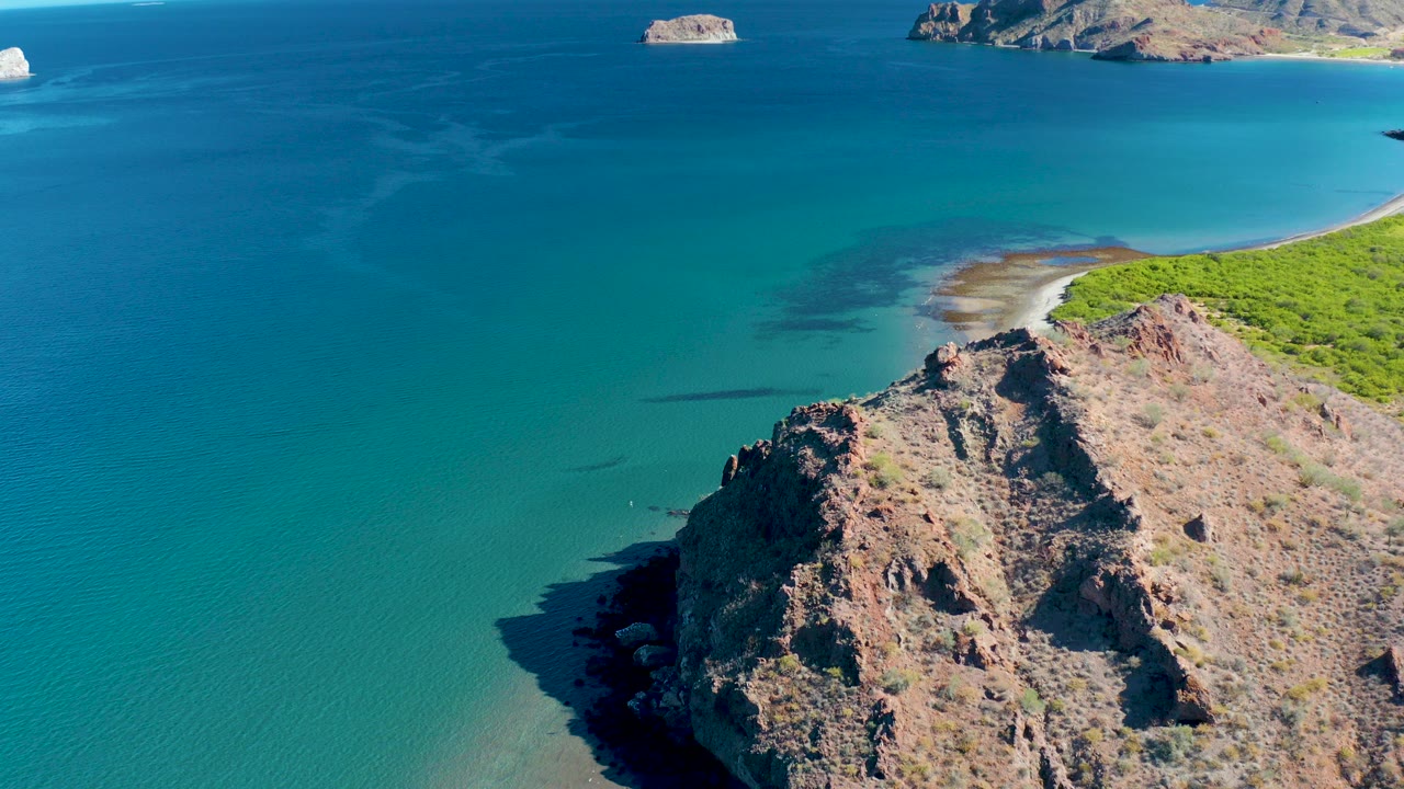 Elevated view in Ligui, Baja California Sur of a rocky cliff above turquoise water, a small sandy shoreline, green coastal vegetation, distant rocky islands, and no visible people.