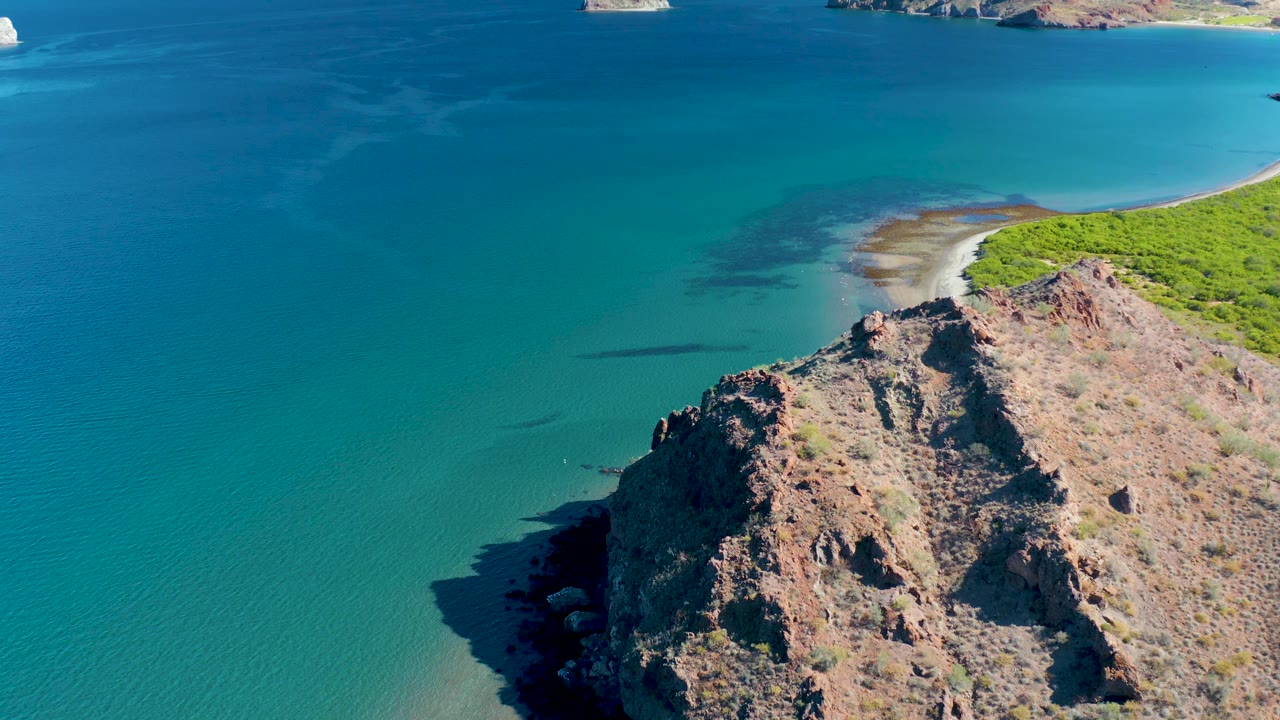 Elevated Ligui coastline view with textured brown rock in the foreground, calm turquoise water to the left, a curving sandy beach at upper right, green vegetation behind it, and no visible people.