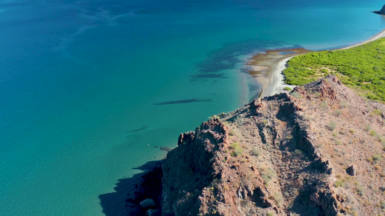 Aerial view in Ligui, Baja California Sur, showing the cliff slightly farther right, a broad field of turquoise water, the sandy shoreline curving behind the rocks, green vegetation at the upper right, and no visible people.