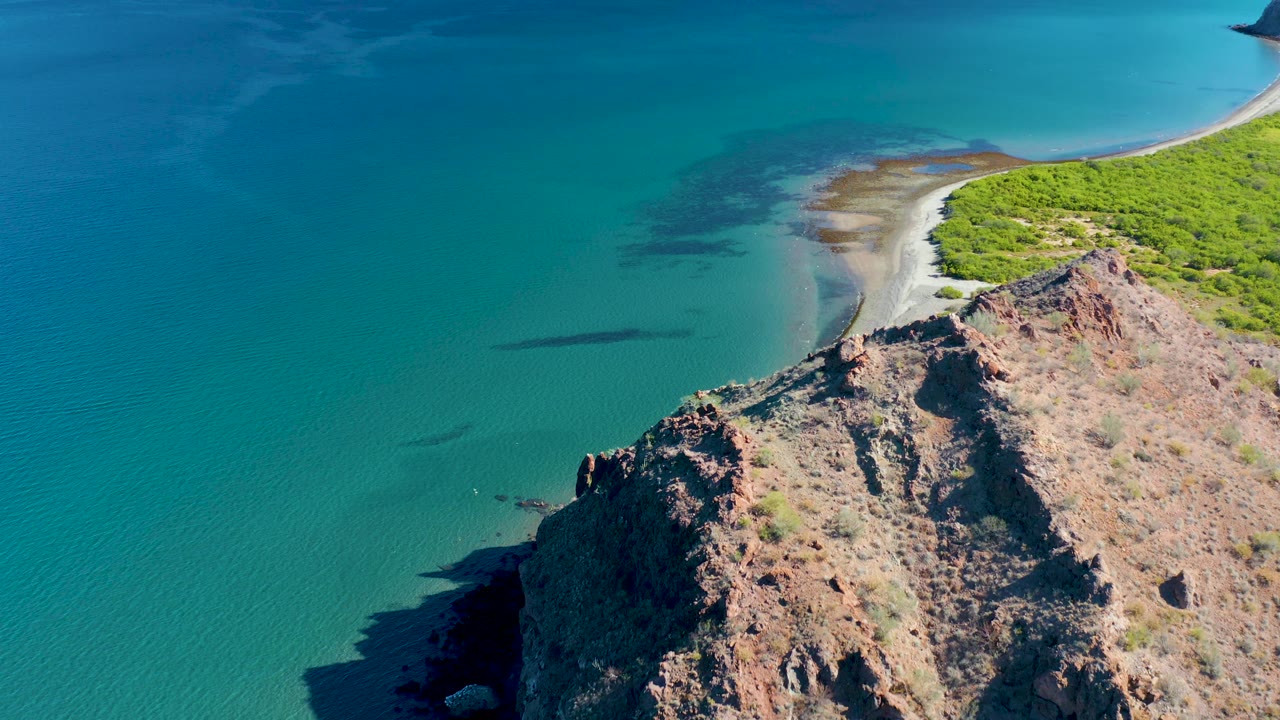 Aerial frame from Ligui, Baja California Sur, with textured brown rock in the lower right, clear green-blue water across the left side, a thin sandy beach at the top, coastal vegetation, and no visible people.