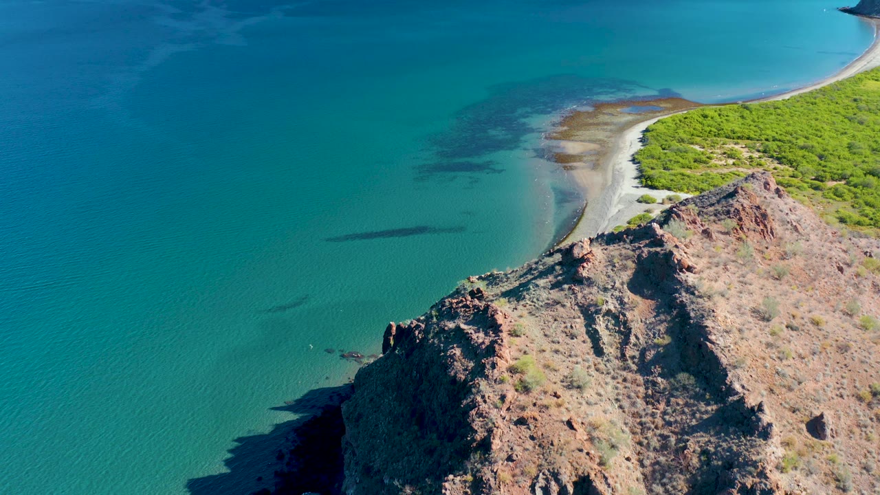 Mid-clip aerial view over Ligui, Baja California Sur, with the cliff edge lower in frame, turquoise water dominating the left half, pale sand and green coastal vegetation along the upper shoreline, and no visible people.
