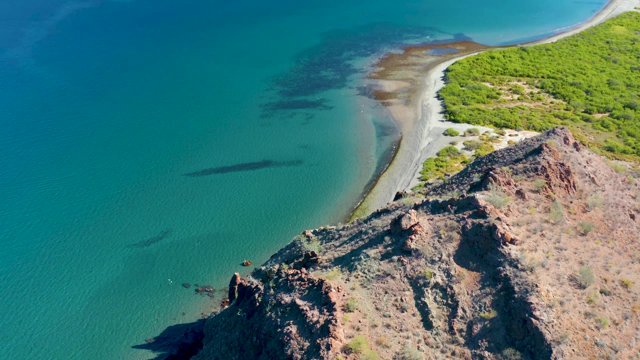 Aerial view in Ligui, Baja California Sur with the rocky point lower in frame, turquoise water to the left, the curved sand beach above it, inland green brush, and no visible people.