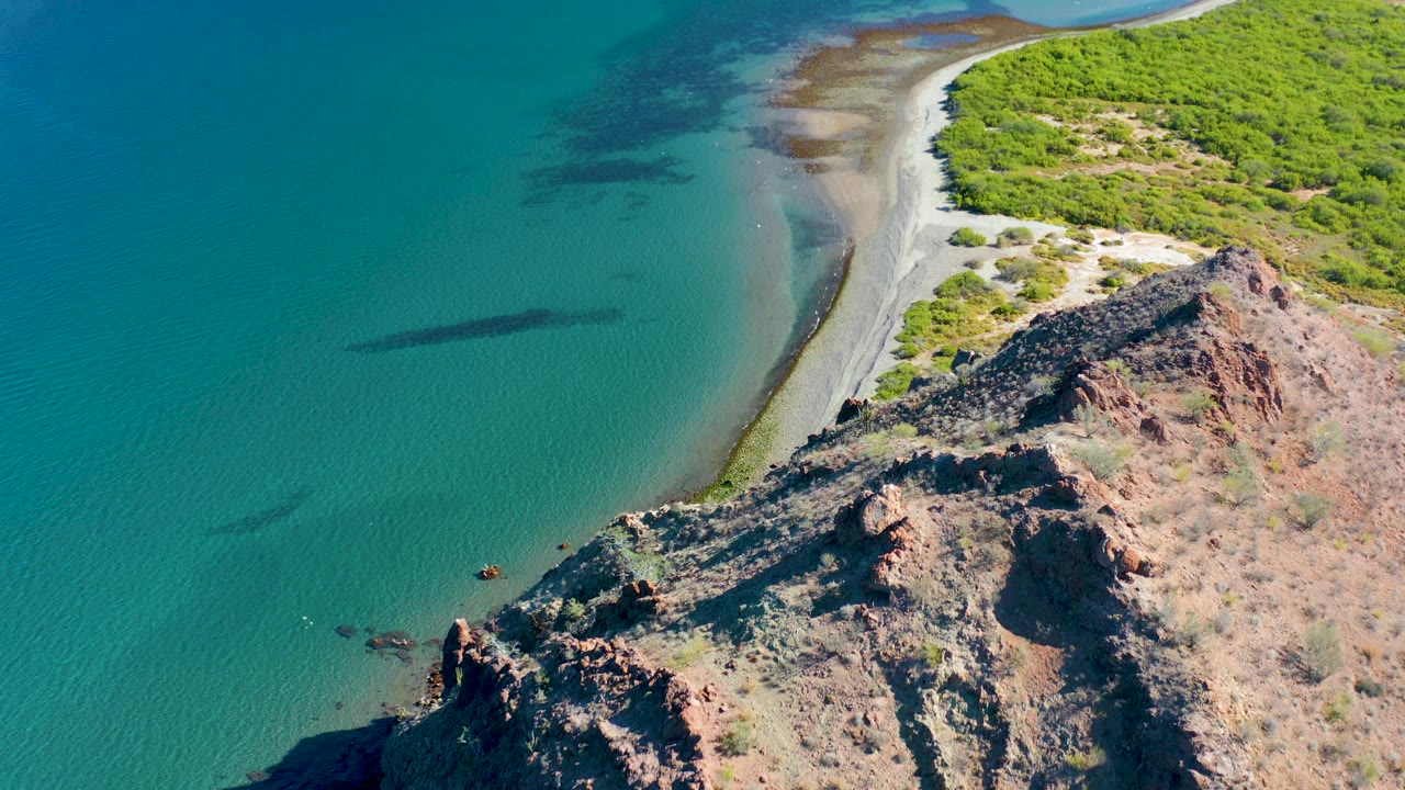 Aerial frame from Ligui, Baja California Sur with turquoise water on the left, a thin pale shoreline through the center, rugged rock in the foreground, scrub-covered coast at top right, and no visible people.