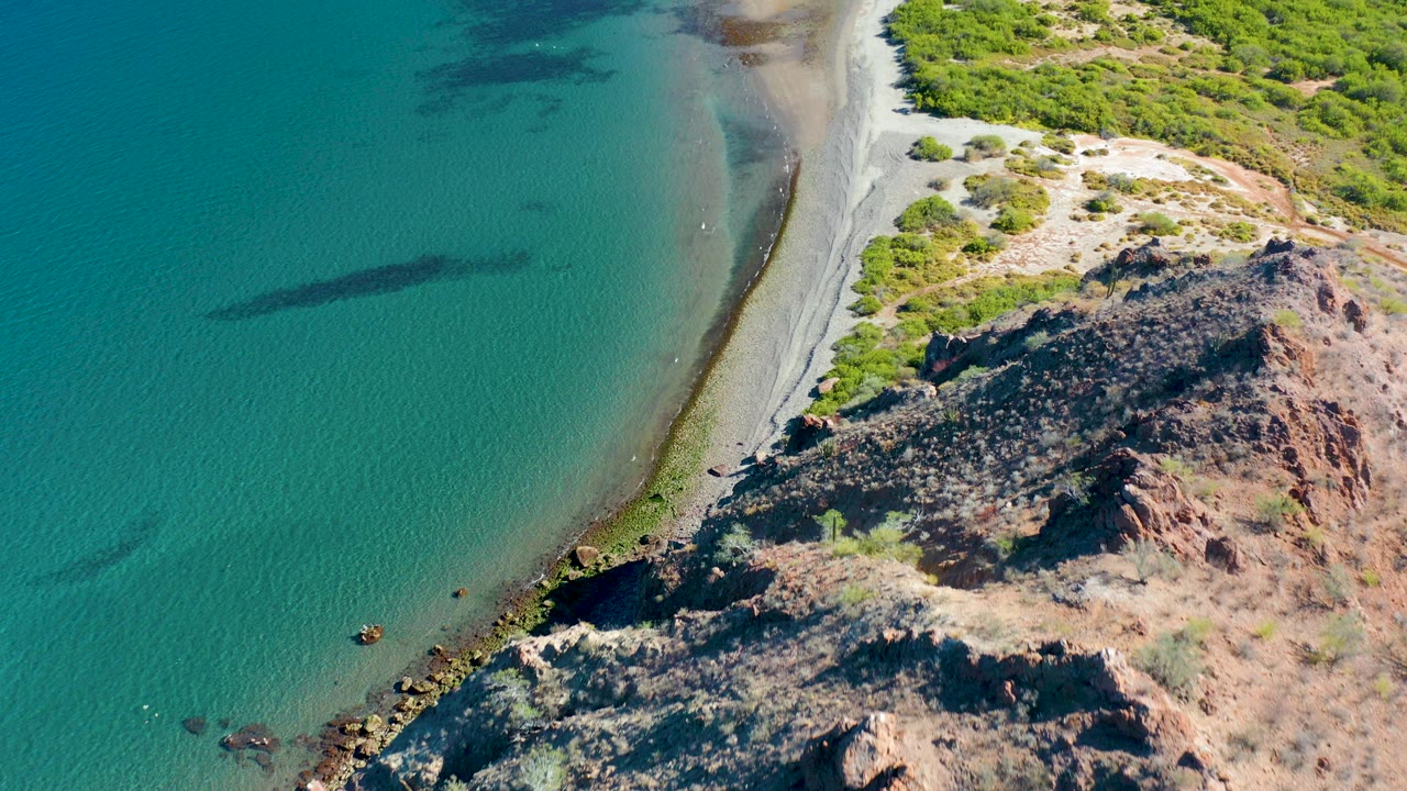 Aerial frame over Ligui, Baja California Sur shows turquoise water beside a pale curving beach, dark rocky outcrops, and green shoreline vegetation, with no visible people.